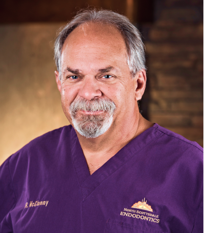 Male dental professional in purple scrubs smiling at camera in clinical setting.