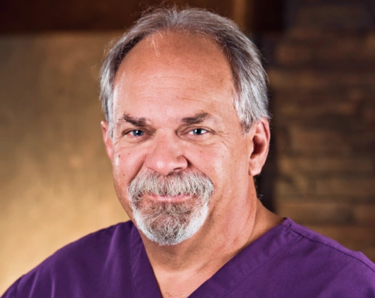 Professional headshot of male dentist in purple scrubs against warm, blurred clinical background.