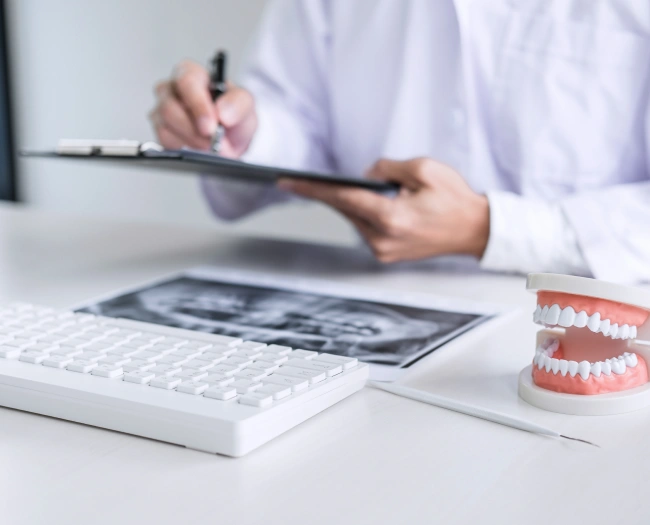 Dentist writing on a clipboard at a desk with a keyboard, dental x-ray, and teeth model.